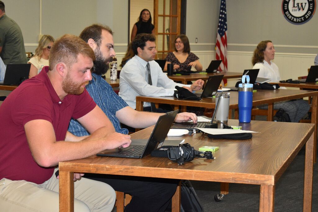 Group of people sitting at tables with laptops and water bottles, a flag in the background.