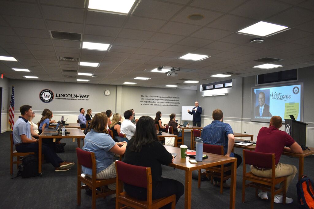 Group of seated individuals in a classroom with tables, a projector screen, and a standing person presenting.