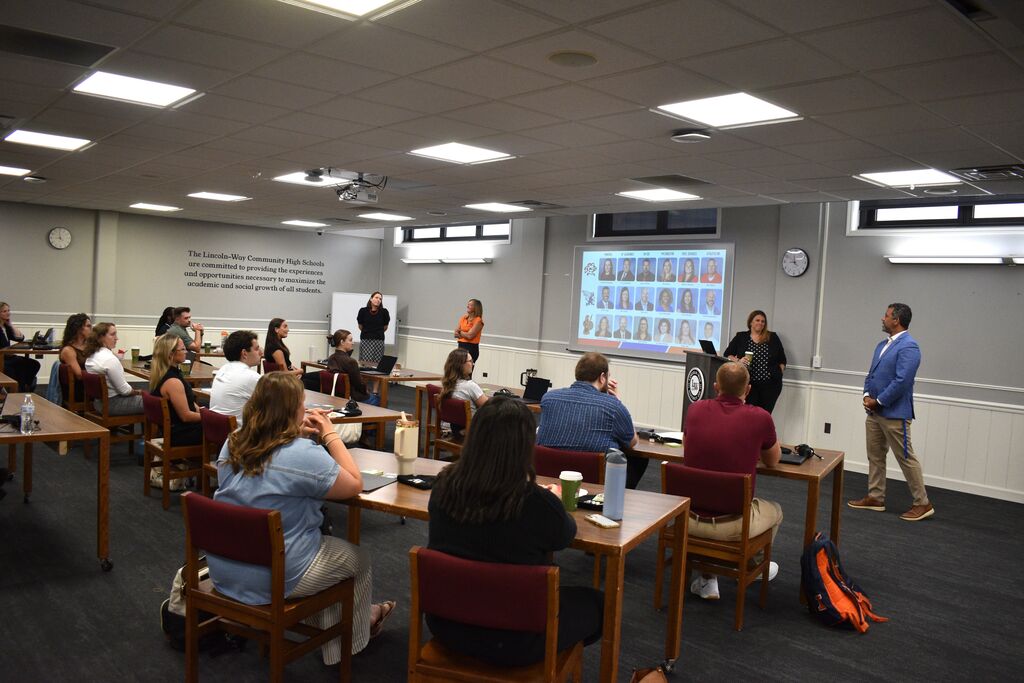 A classroom with a presentation in progress. Participants are seated at tables, listening. A person stands at the front, addressing the group.