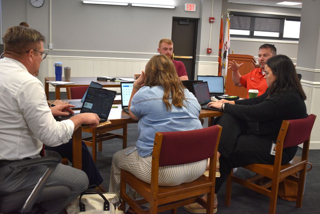 Six people seated around a table, each using a laptop. A clock is on the wall, and a door is behind them.