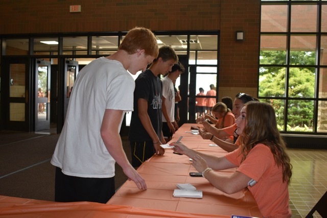 Several people sit at a long orange table with various items. A woman hands a paper to a man.