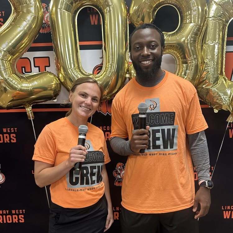 Two individuals, a man and a woman, stand together in front of balloons reading "2020." They wear matching orange shirts.