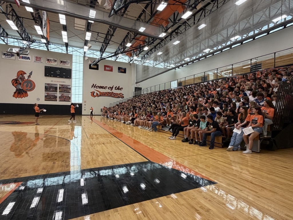 Large indoor basketball court with a crowd of people sitting on bleachers. The court has black and orange markings.