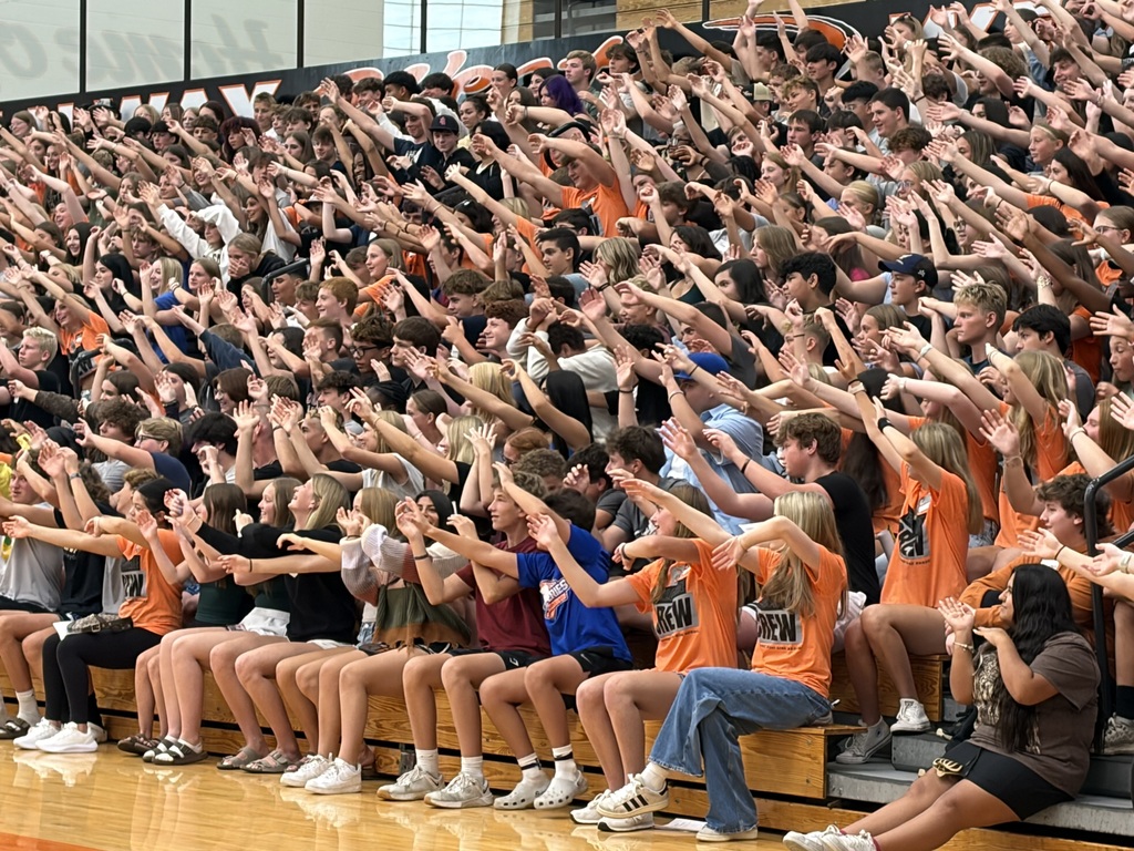 A large crowd of people wearing orange shirts seated on bleachers, raising their hands in a school gym.