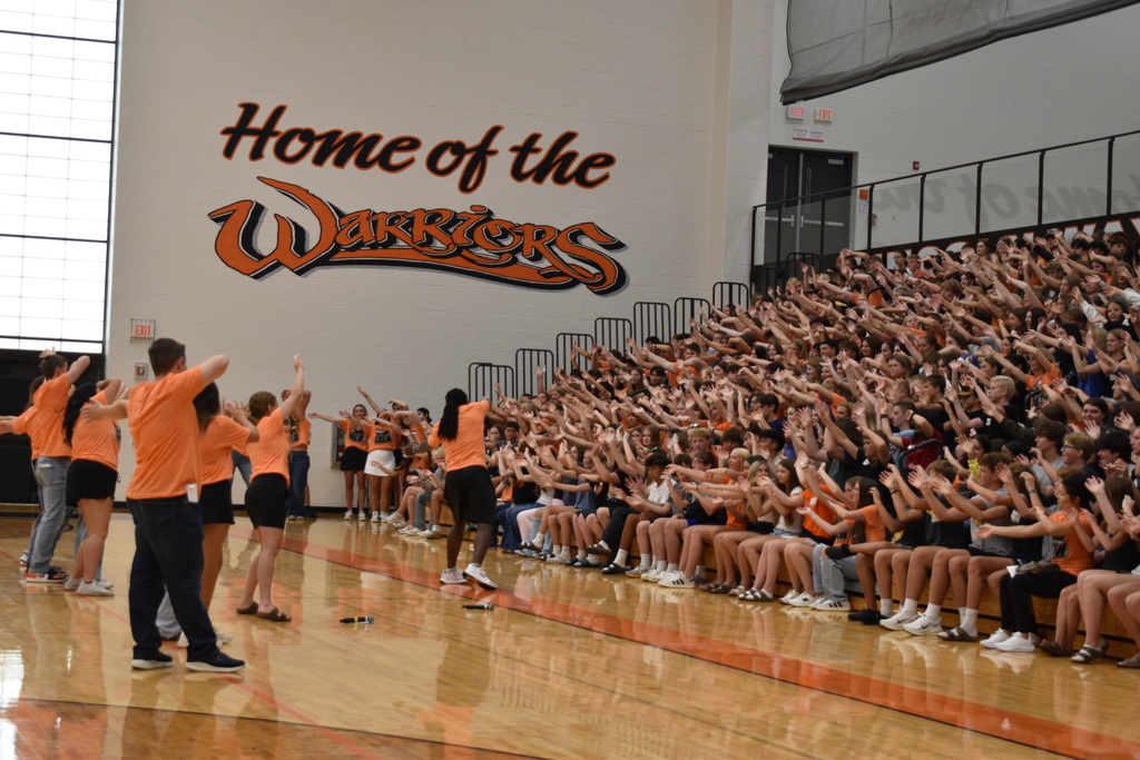 An indoor basketball court with a crowd of people, some standing, cheering, and holding hands, while others sit on bleachers.