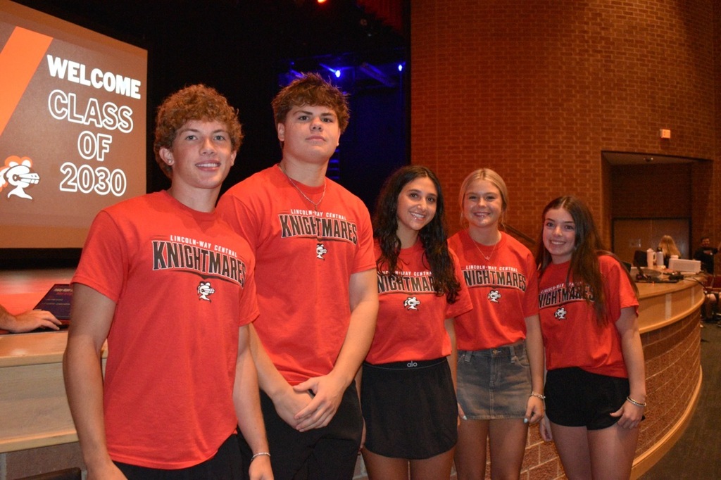 Five young adults, all wearing orange shirts, stand close together in front of a large screen.