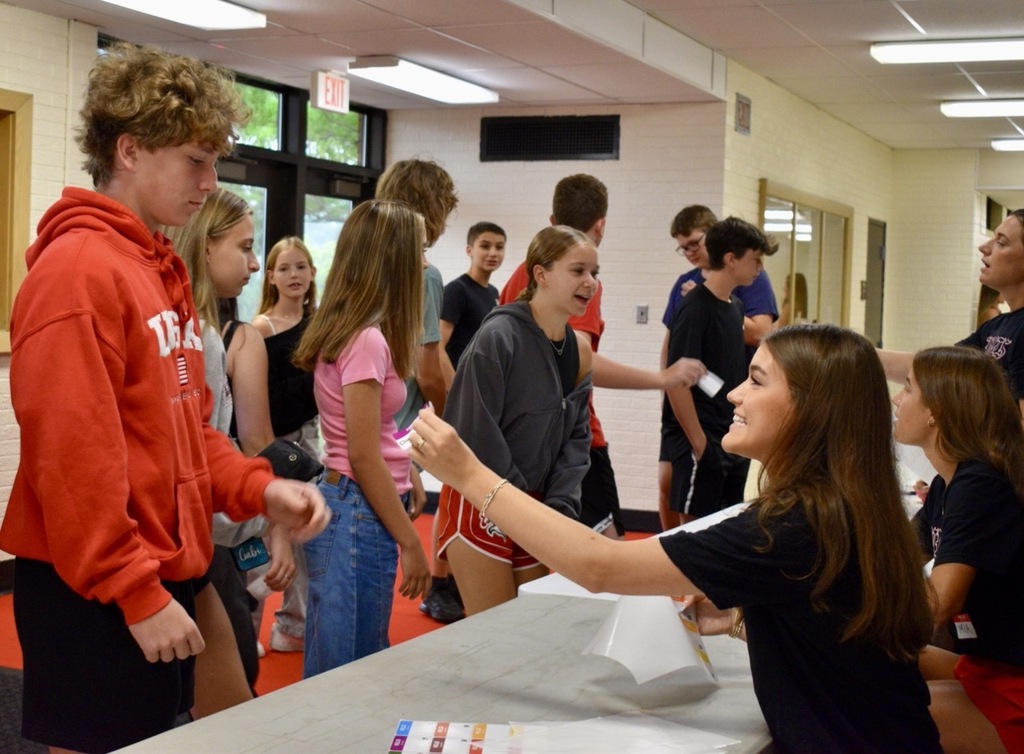 Teens in a school hallway interact with a woman at a table. Some students wear hoodies and sneakers.