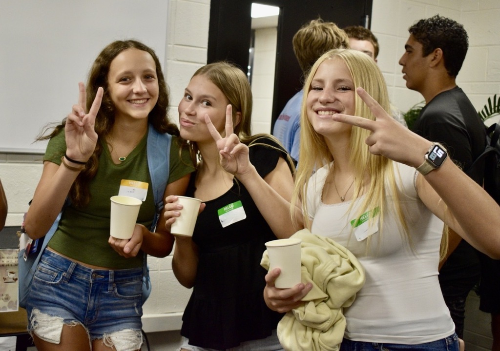 Three women make peace signs and point fingers in front of a white wall. Two hold white cups.