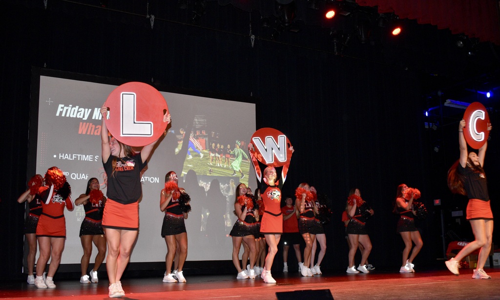 Group of cheerleaders holding large letters L and C on stage, projector screen behind them showing "Friday".