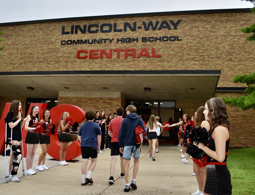 Students wearing school colors stand in front of a building with a sign reading "LINCOLN-WAY COMMUNITY HIGH SCHOOL CENTRAL".