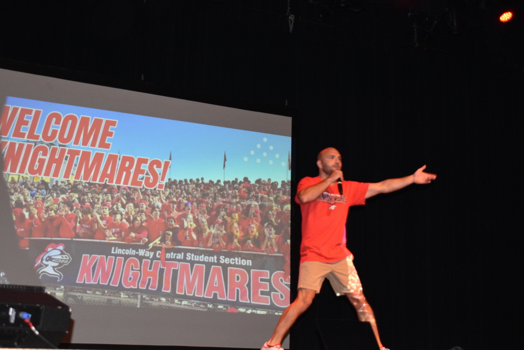 Person speaking on a stage, wearing red shirt and shorts. Behind him, a large screen displays "Welcome Nightmares!"