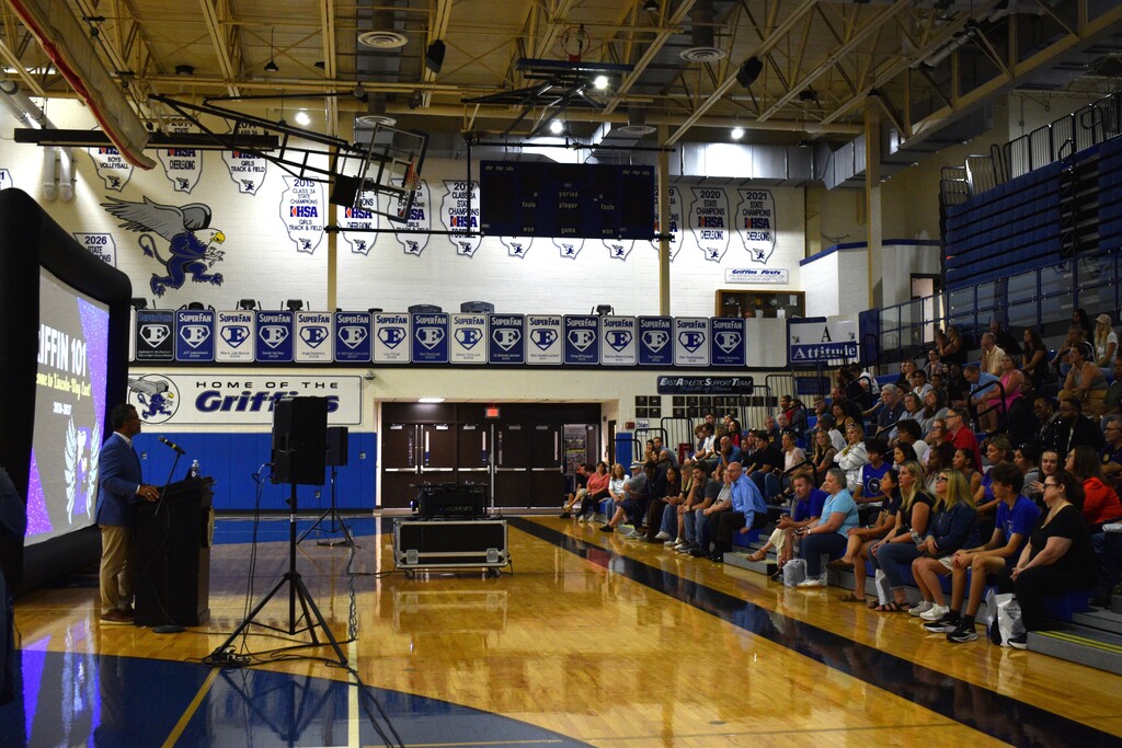 A man stands at a podium on a basketball court, addressing a seated crowd with a projector screen behind him.