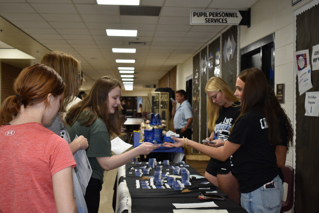 People are standing around a table filled with items, handing out items to two other women.