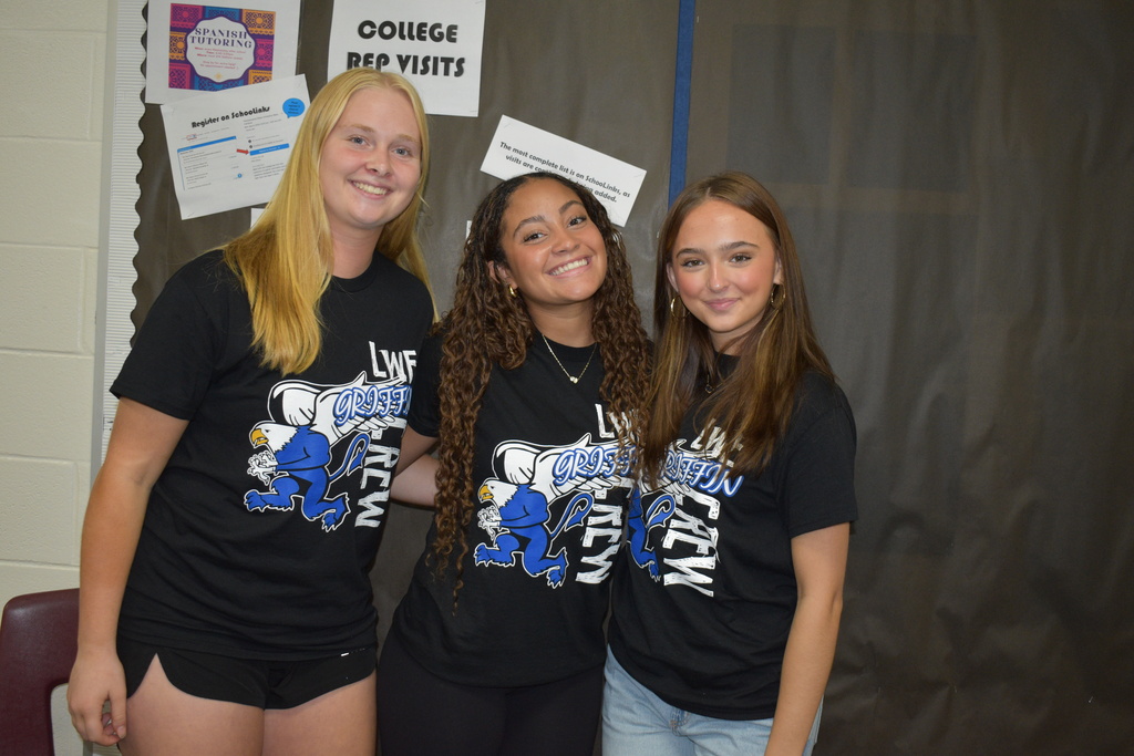 Three students stand next to each other, wearing identical black T-shirts. Background has signs related to college.
