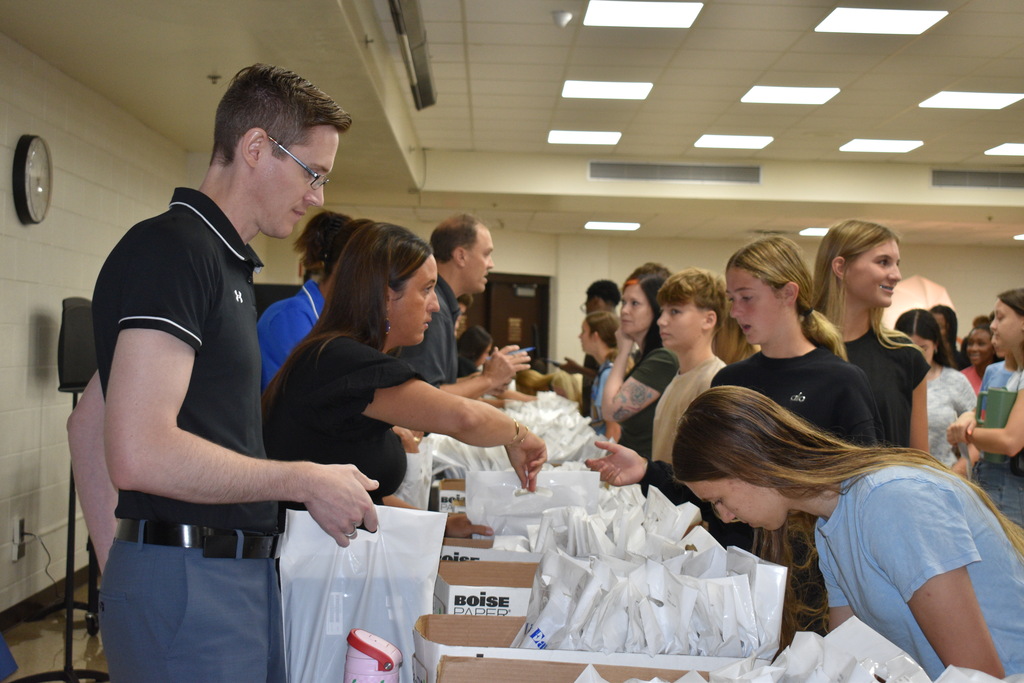 People stand at a table with bags and boxes, one man holds a bag, another leans over to hand out items.