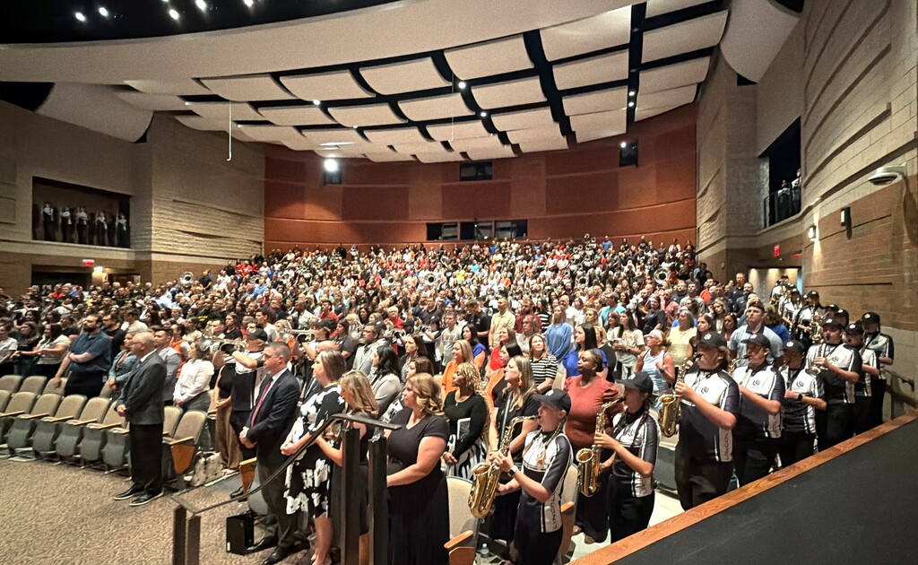 Large audience in an auditorium watching musicians play instruments on a stage. Chairs fill the audience area.