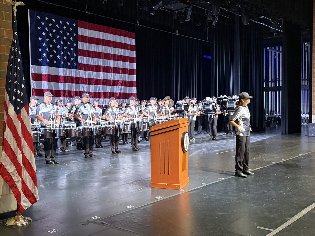 A person stands at a podium with an American flag on stage; a marching band plays behind them.