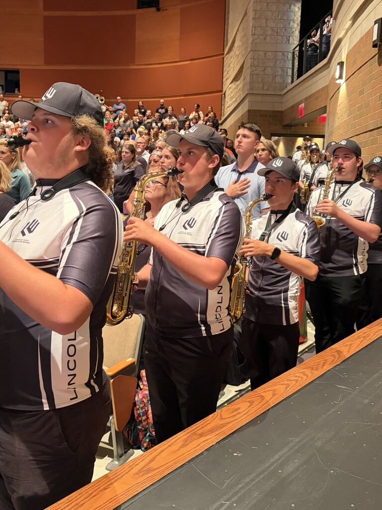 A group of musicians wearing matching black and white uniforms with "Lincoln Way" on the back, playing saxophones in a filled auditorium.