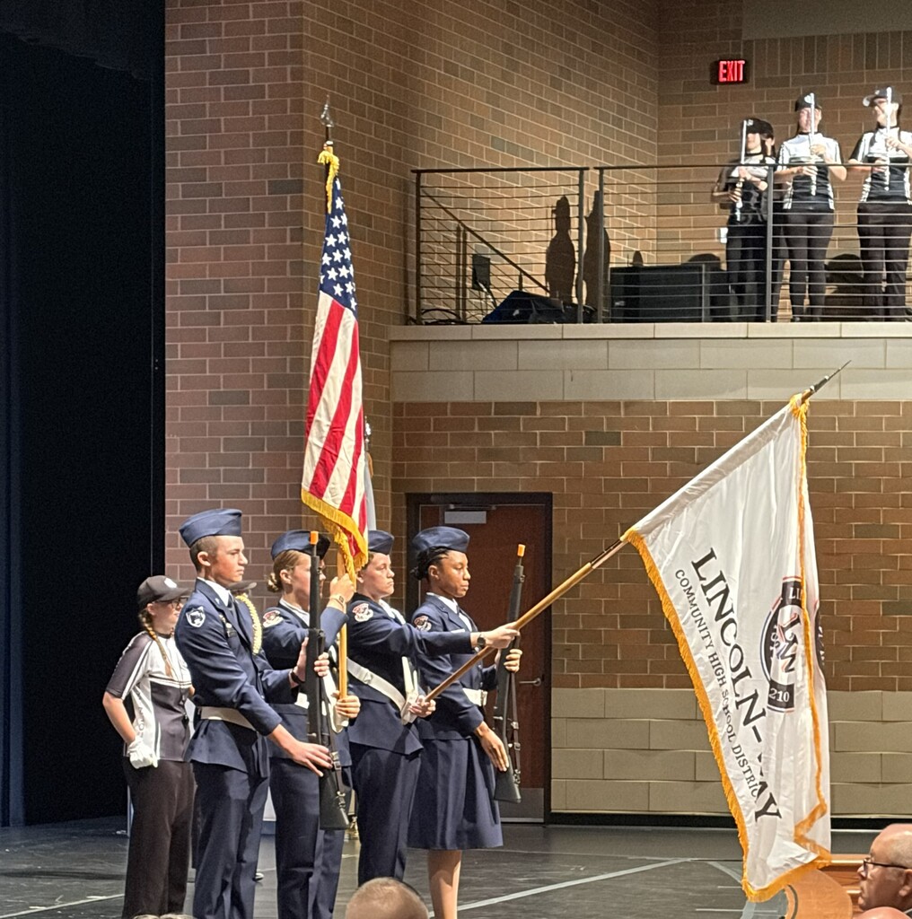 Uniformed people on stage hold flags. An American flag stands on the left. Behind them, a group stands on a balcony.