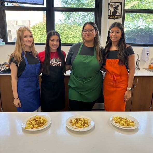 Four women wearing aprons in a kitchen. Three plates of food on a counter. Windows and trees in the background.