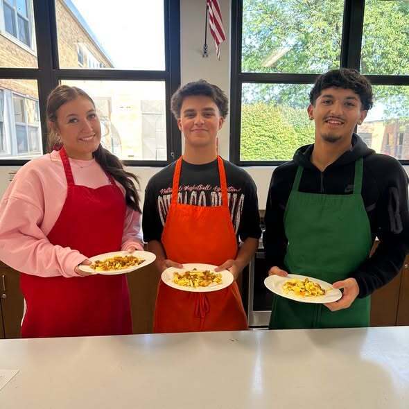 Three individuals in aprons, holding plates of food, standing by a kitchen counter with a window and flag in the background.