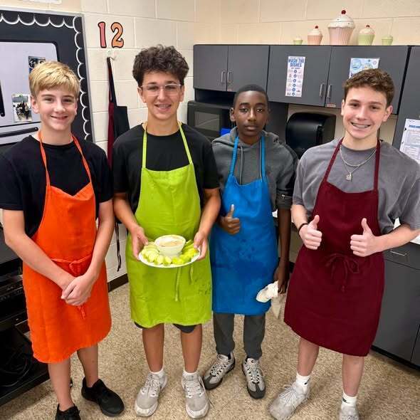 Four young men in aprons stand in a kitchen. One holds a dish with food.