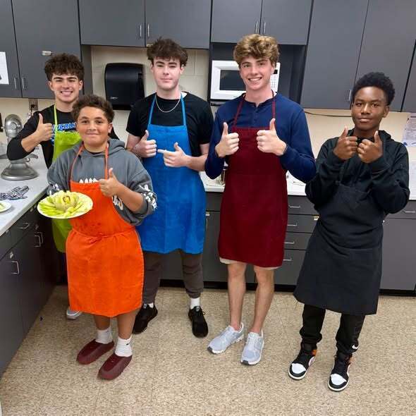 Five individuals in aprons, likely students, pose in a kitchen with gray cabinets and a tiled floor.