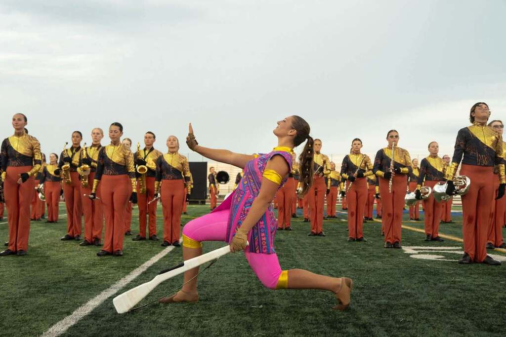 Person in pink and purple uniform performing on field, with band in red and black uniforms behind her.