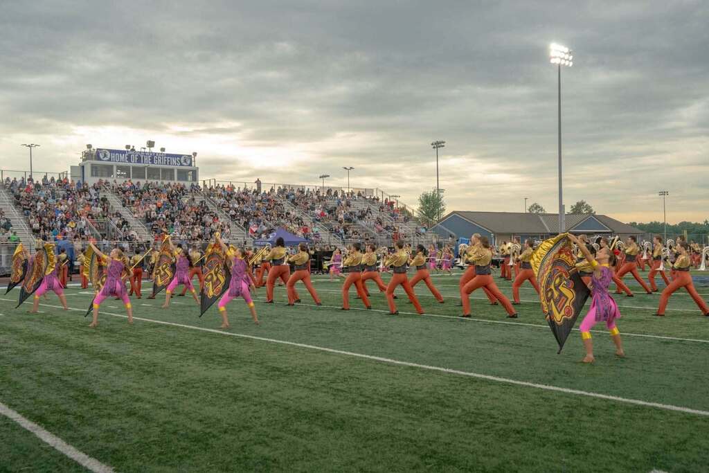 A marching band performs on a field at dusk. Audience in stands. Bright stadium lights. Cloudy sky.