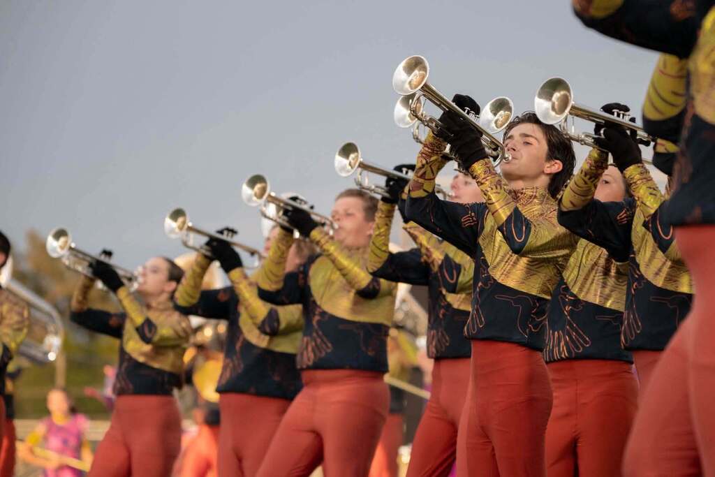 A group of individuals in black and gold uniforms playing trumpets on a field with a cloudy sky.