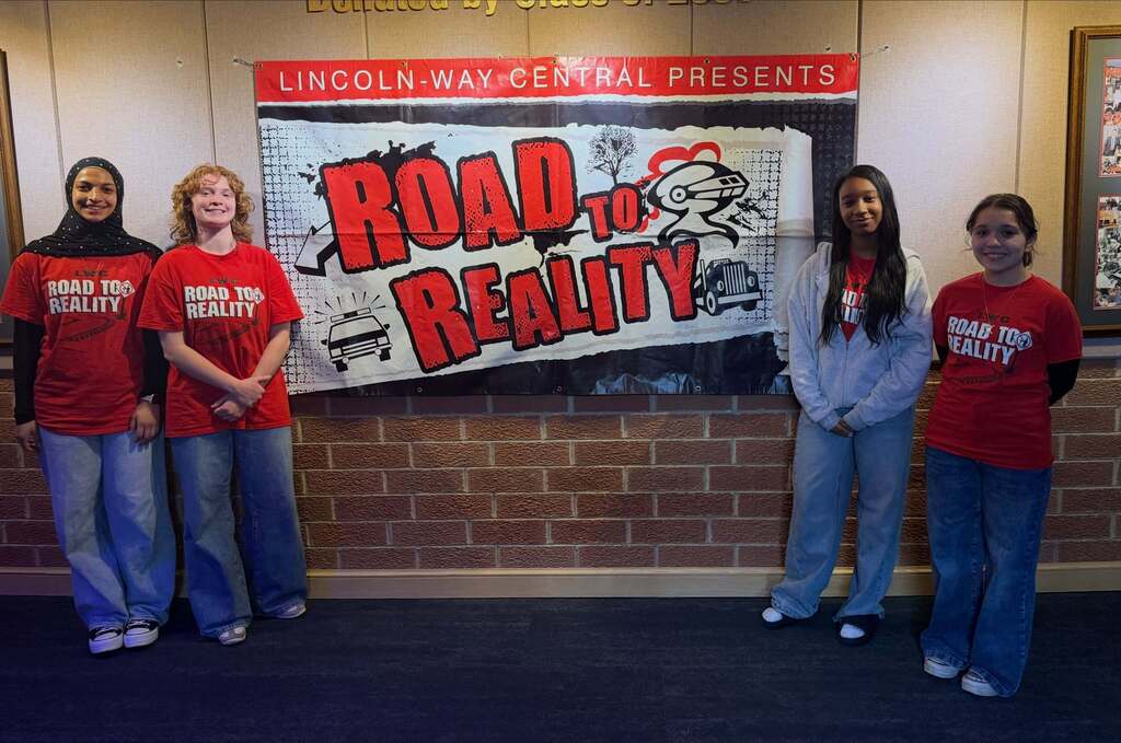 Five young people stand in front of a brick wall with a "Road to Reality" banner.