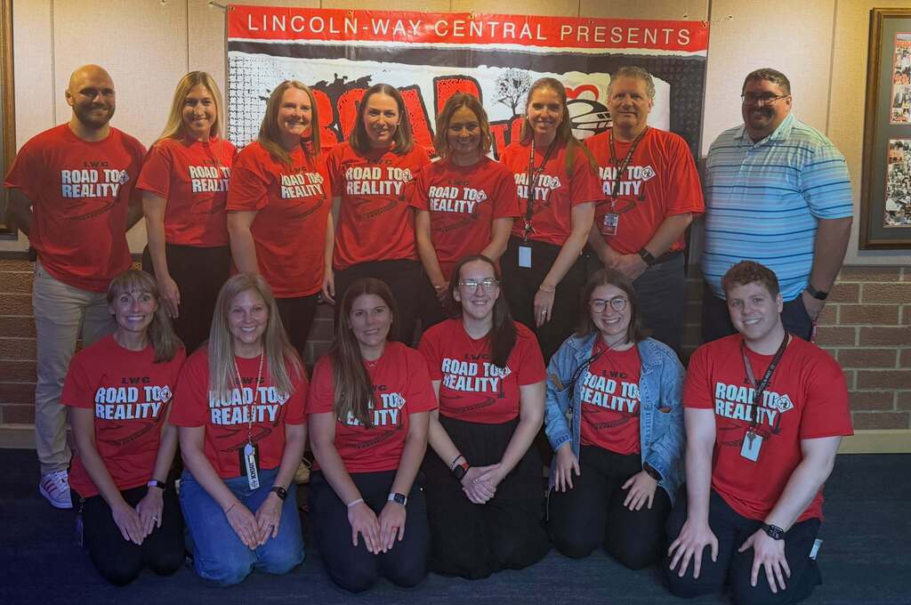 Group of people wearing red shirts with white text, posing for a photo in front of a banner.