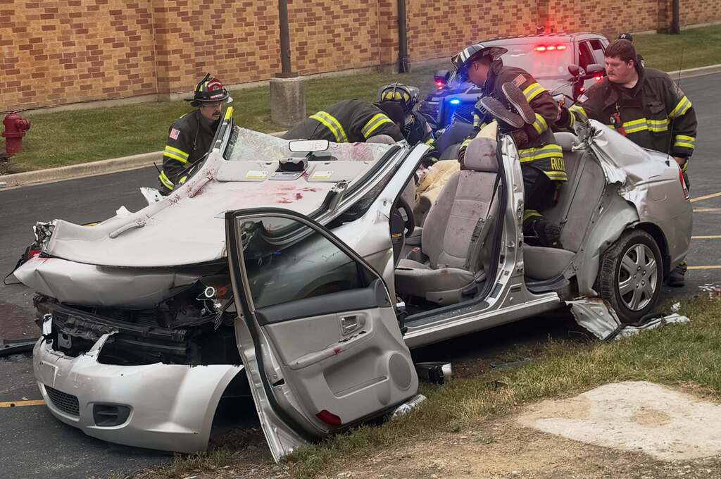 Firefighters at a car crash site, with a damaged silver vehicle. Doors open, engine exposed.