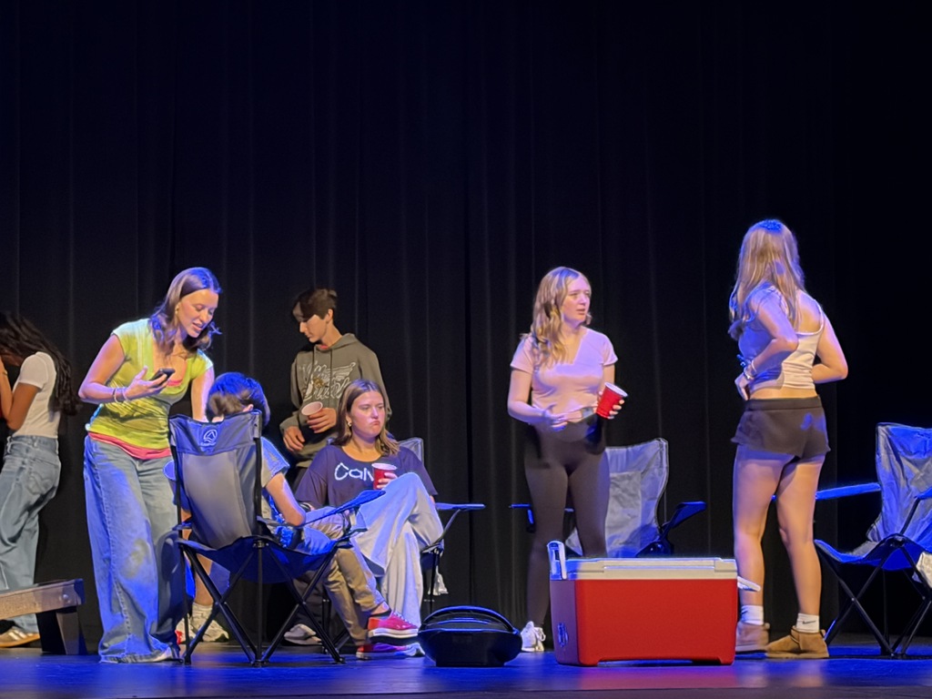 On a stage with a black backdrop, eight individuals stand, sit, and interact. Some hold cups or food.