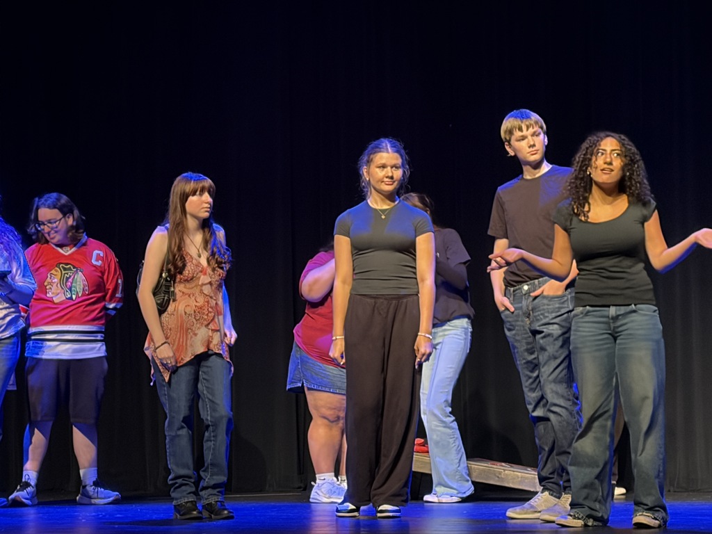 A group of people, some in casual clothing, stand on a stage with a blue backdrop.