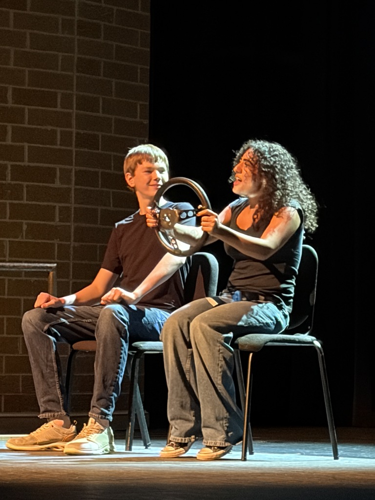 Two people sit on stage chairs; one holds a steering wheel while the other leans forward. Behind them, a brick wall.