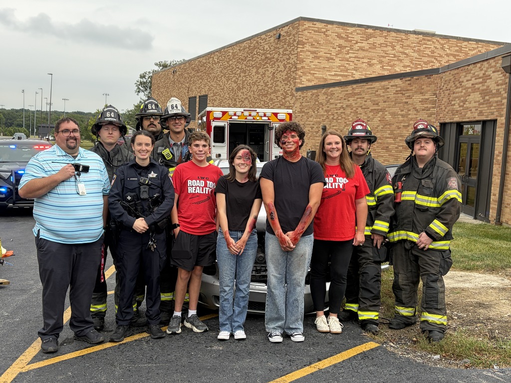 People stand in a parking lot in front of a building; some wear red T-shirts, while others wear firefighter uniforms.