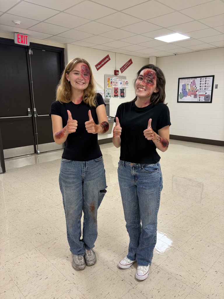 Two individuals stand in a hallway, giving thumbs-up. Both wear black t-shirts and jeans with fake blood on their faces.