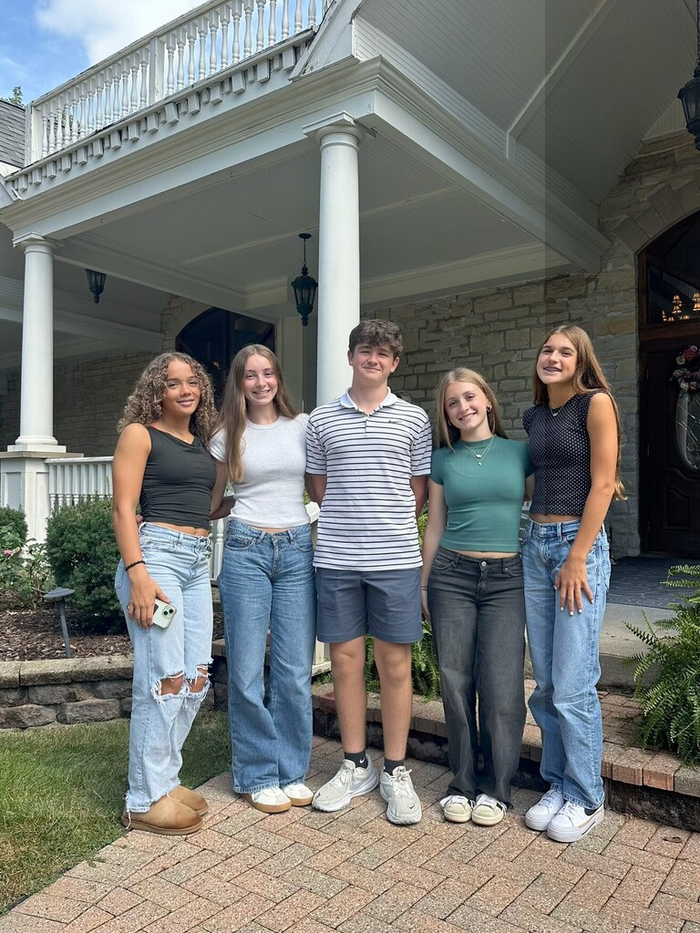 Five people stand in front of a house with a porch. They wear casual clothes and sneakers. Two people on the right wear high-rise jeans.