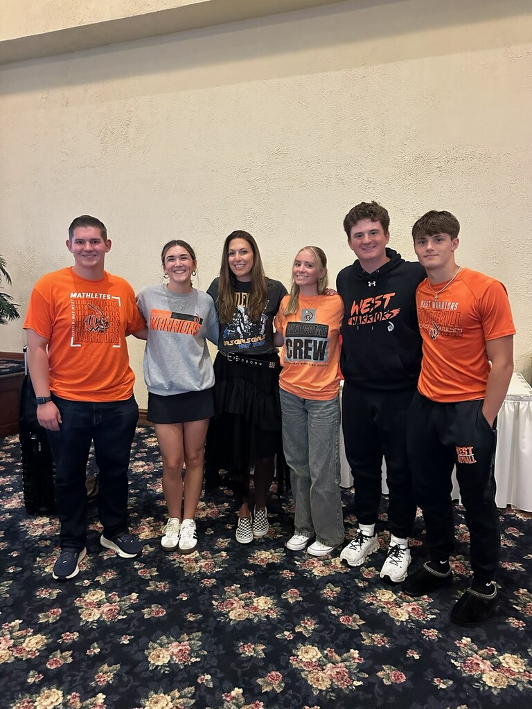 Five individuals in orange and gray tops stand in a room, one holding a trophy. Behind them, a white tablecloth covers a table.