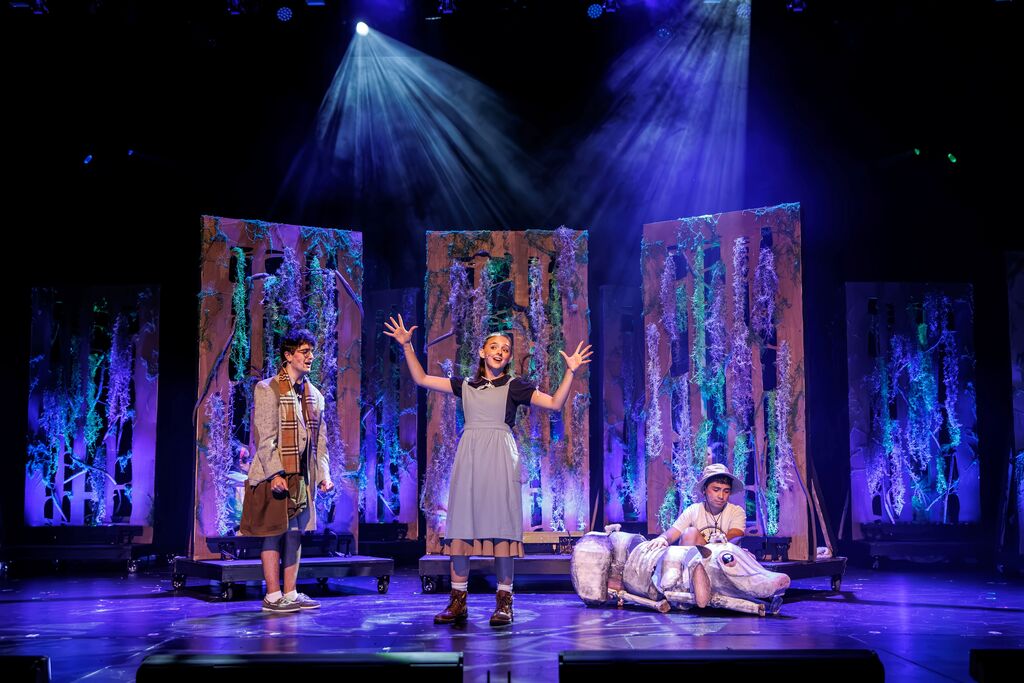 Three actors on stage with one raising hands, another seated, and a backdrop of colorful, textured panels.