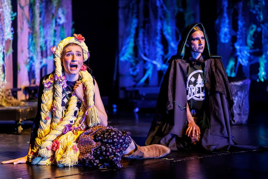 Two actors on stage, one in colorful attire and the other in dark clothing, both seated.