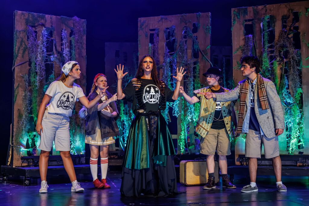 Five people in costumes stand on a stage under spotlights, with two children in front. Green and purple lights illuminate the background.