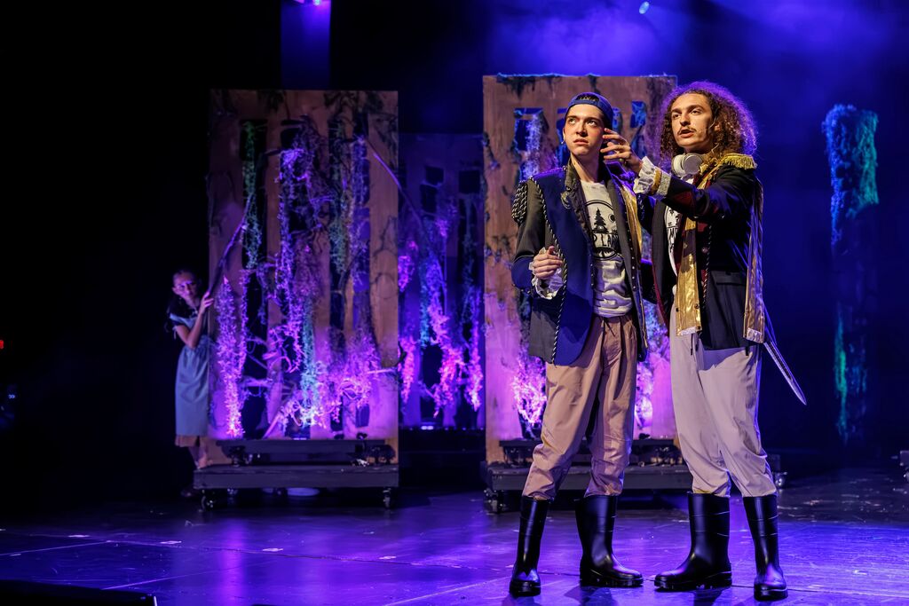 Two individuals on stage, one gesturing, the other holding a sword. Background has abstract figures and purple lighting.