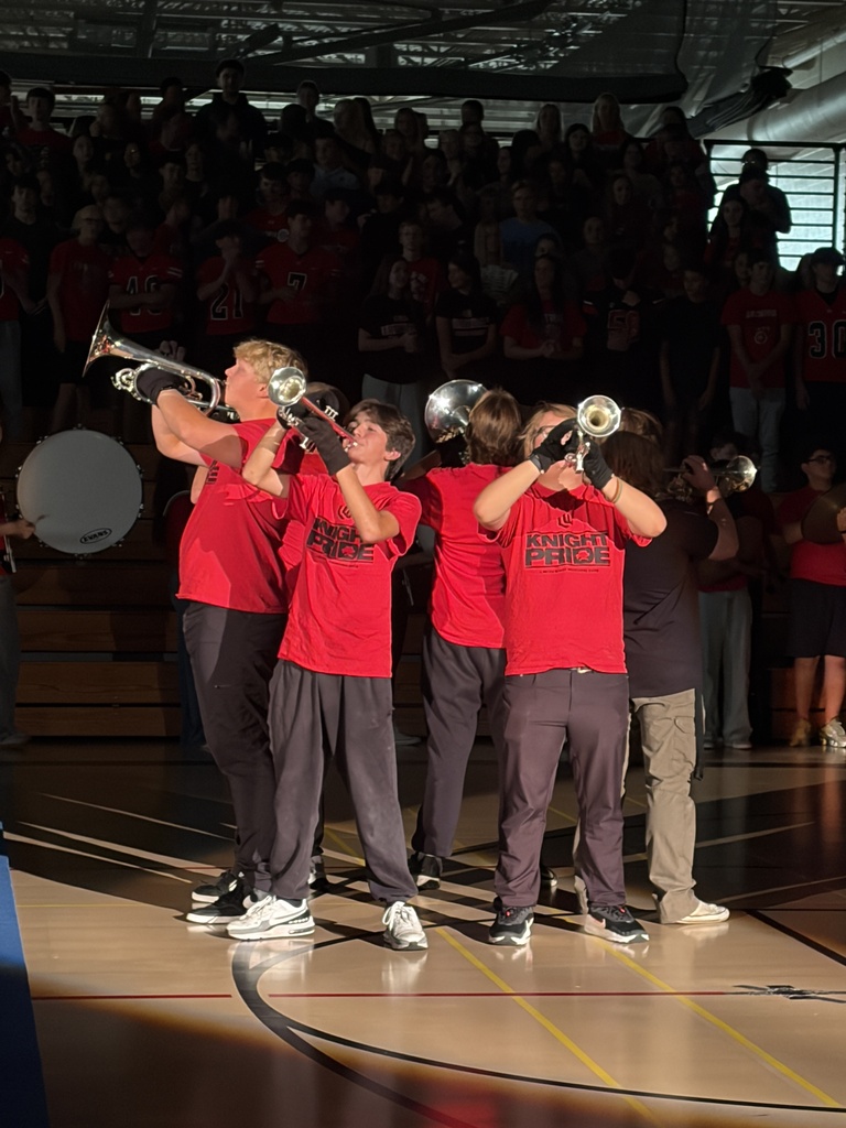 Five students in red shirts and dark pants play trumpets on a basketball court. Audience seated in bleachers in background.