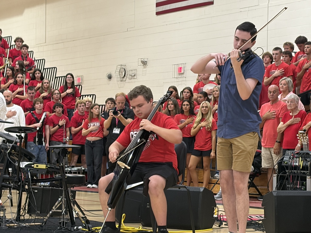Two men play musical instruments on a stage in a gymnasium in front of a cheering audience.