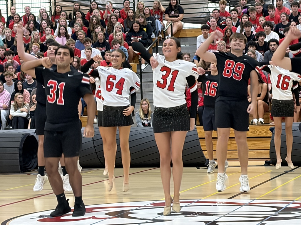 Several cheerleaders on a basketball court, performing in front of a cheering crowd. The cheerleaders are wearing numbered shirts.