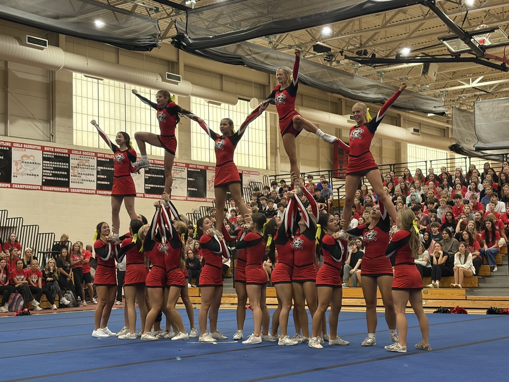 Cheerleaders in red uniforms, performing stunts in a gymnasium with a large audience watching.