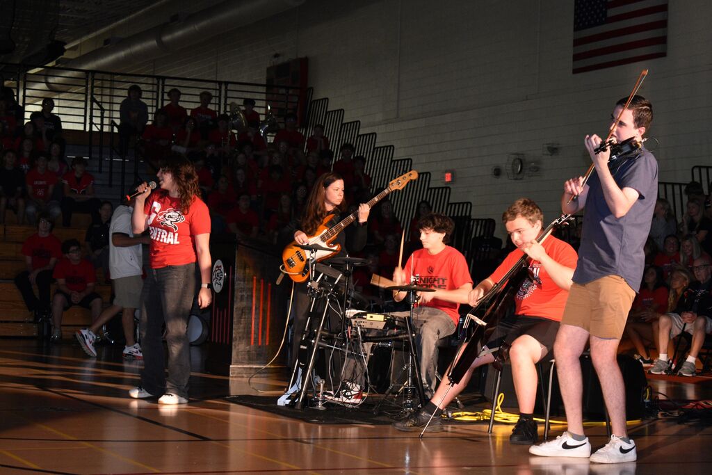 A group of musicians performs on a stage in front of an audience. Four individuals play instruments while another holds a microphone.