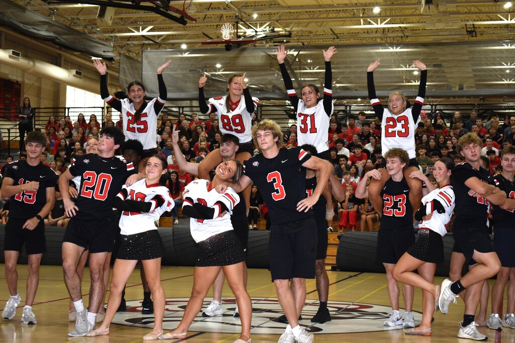 Athletes in black and white uniforms cheer on the court, some standing on shoulders, with a crowd watching.